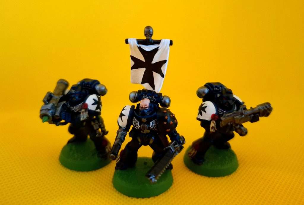 Tres figuras de black templars, al centro un sargento con espada sierra y estandarte, a sus costados marines espaciales con armas especiales, un lanzacohetes y un lanzallamas, respectivamente